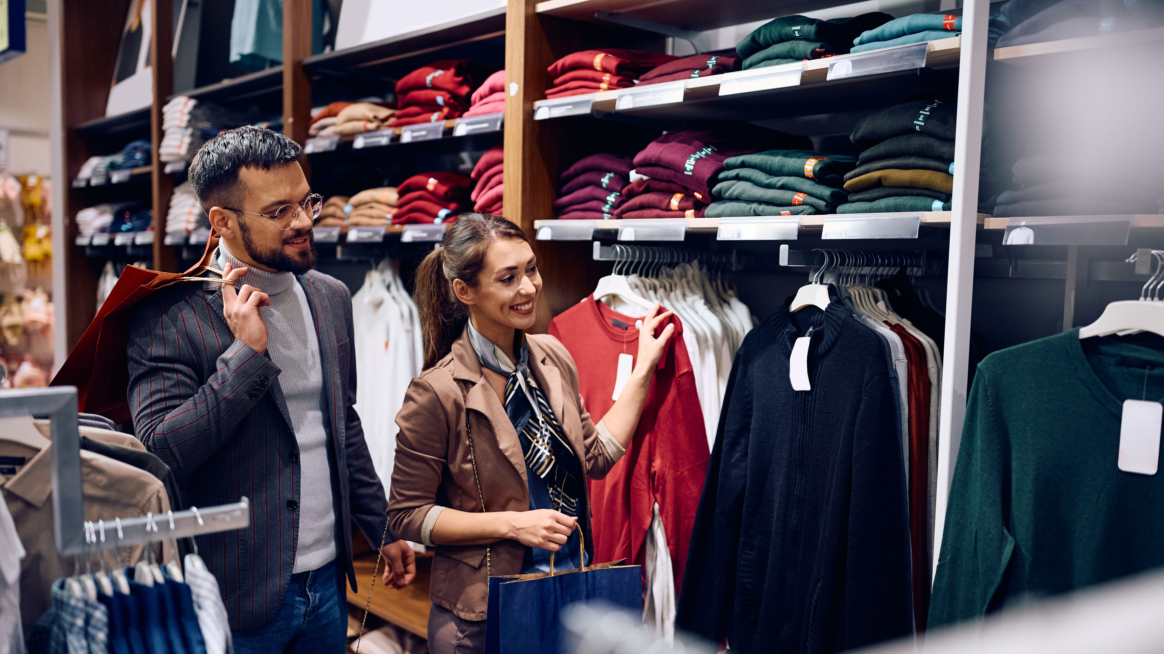 A man and a woman shopping for clothes in a retail store, browsing a rack of sweaters.






