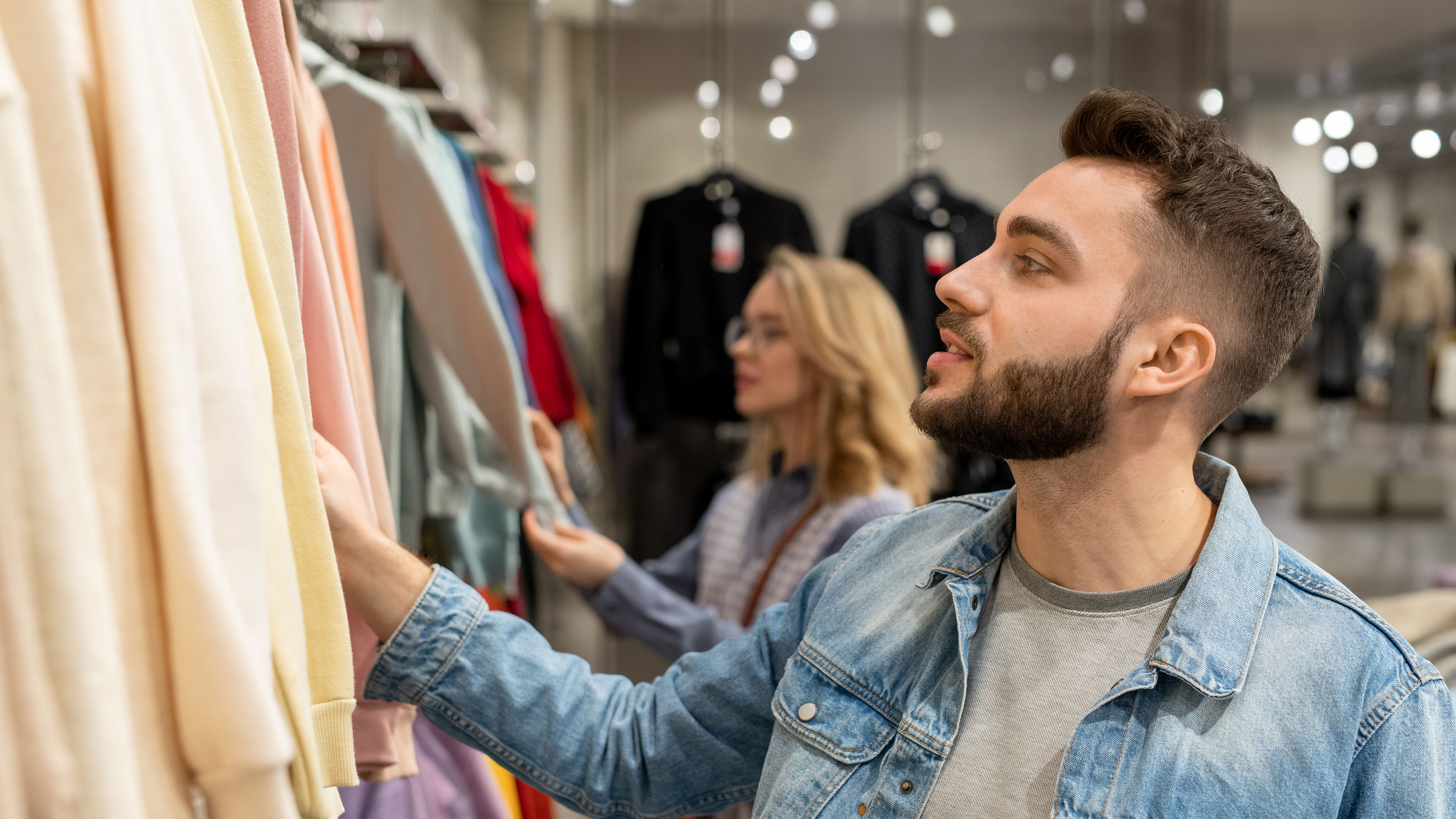 A stylish man browsing clothes in a store, with a slightly blurred woman in the background.